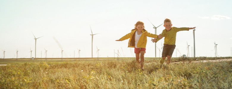 A young girl and boy running through a field with wind turbines in the background